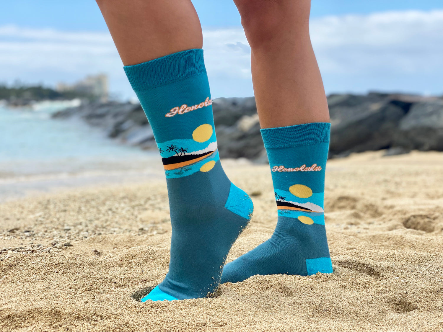 Blue socks with a beach design and the word "Honolulu", worn on a woman's feet on the beach.